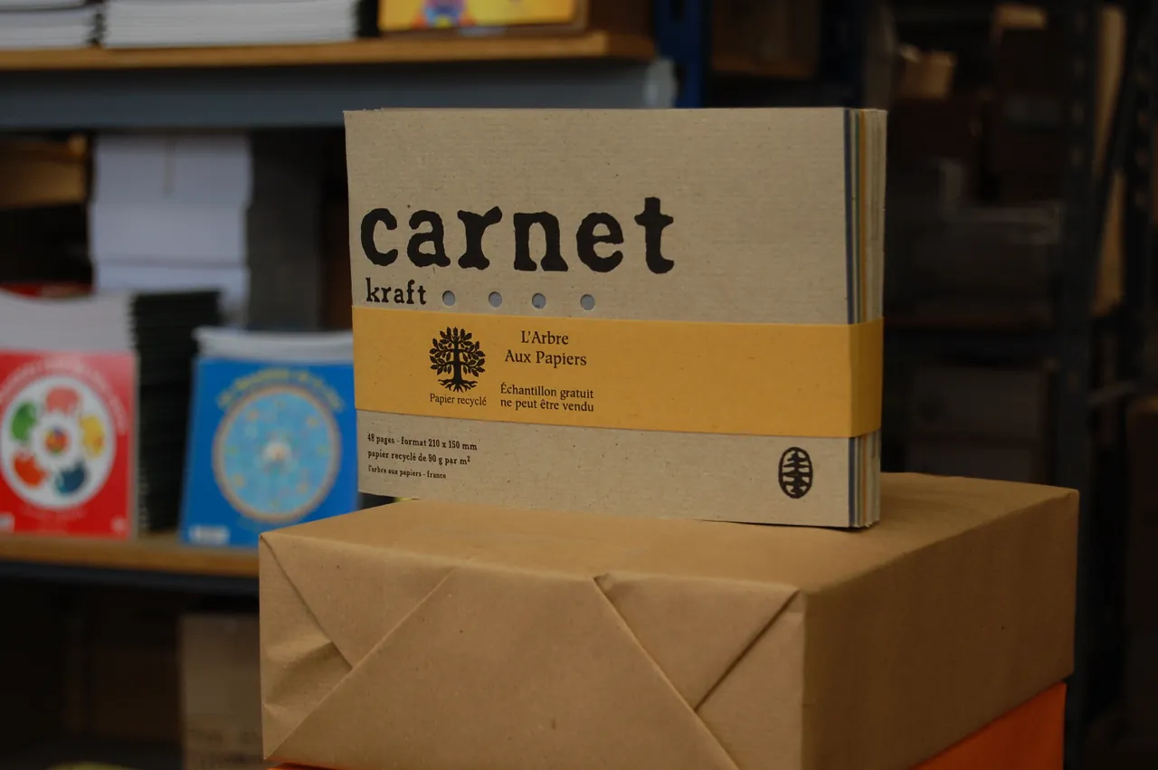 Un carnet kraft de l'Arbre aux Papiers avec son bandeau moutarde, posé sur une rame de papier dans l'atelier